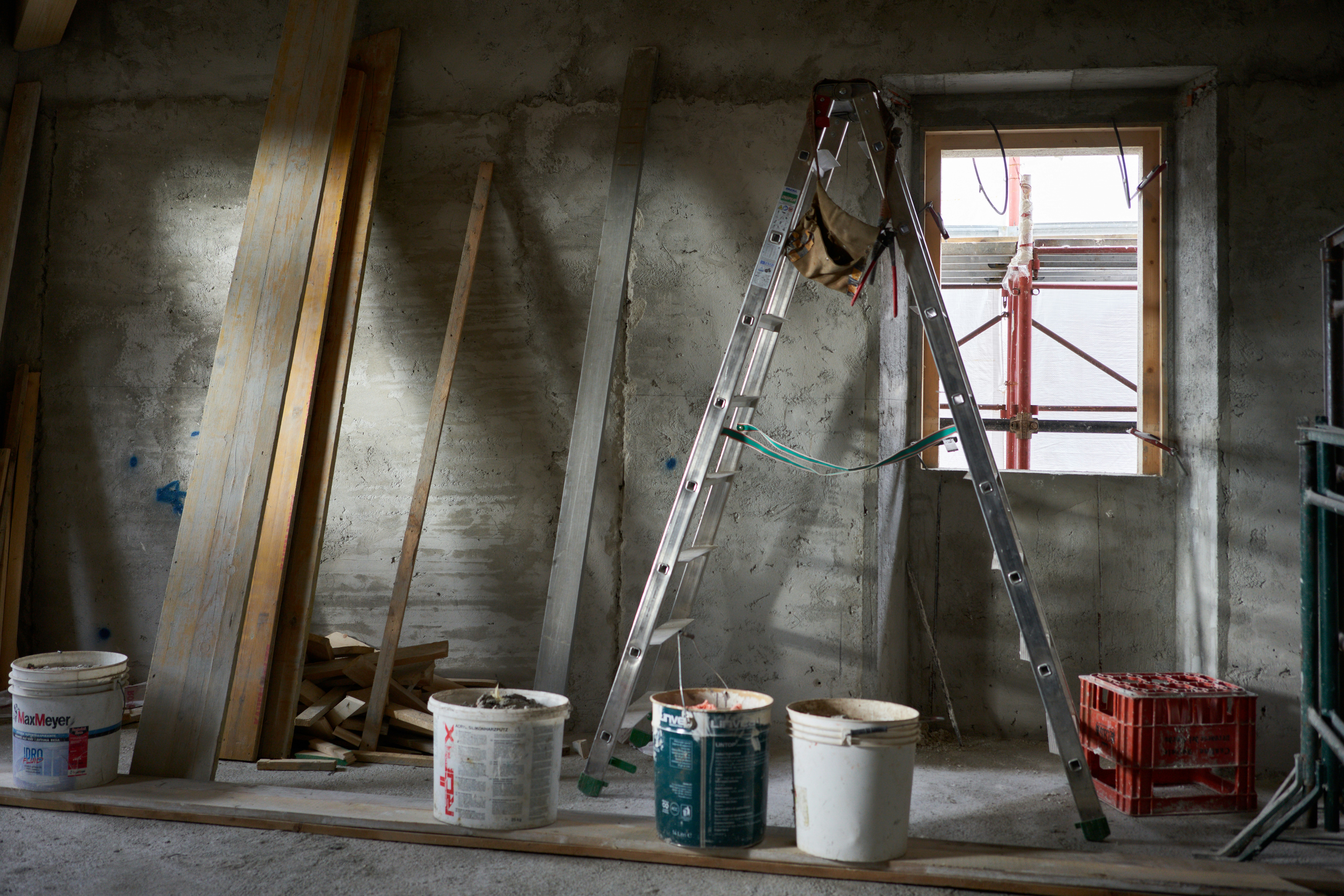 Unfinished renovation site interior with bare concrete walls, a ladder, paint buckets and timber offcuts beside an open window