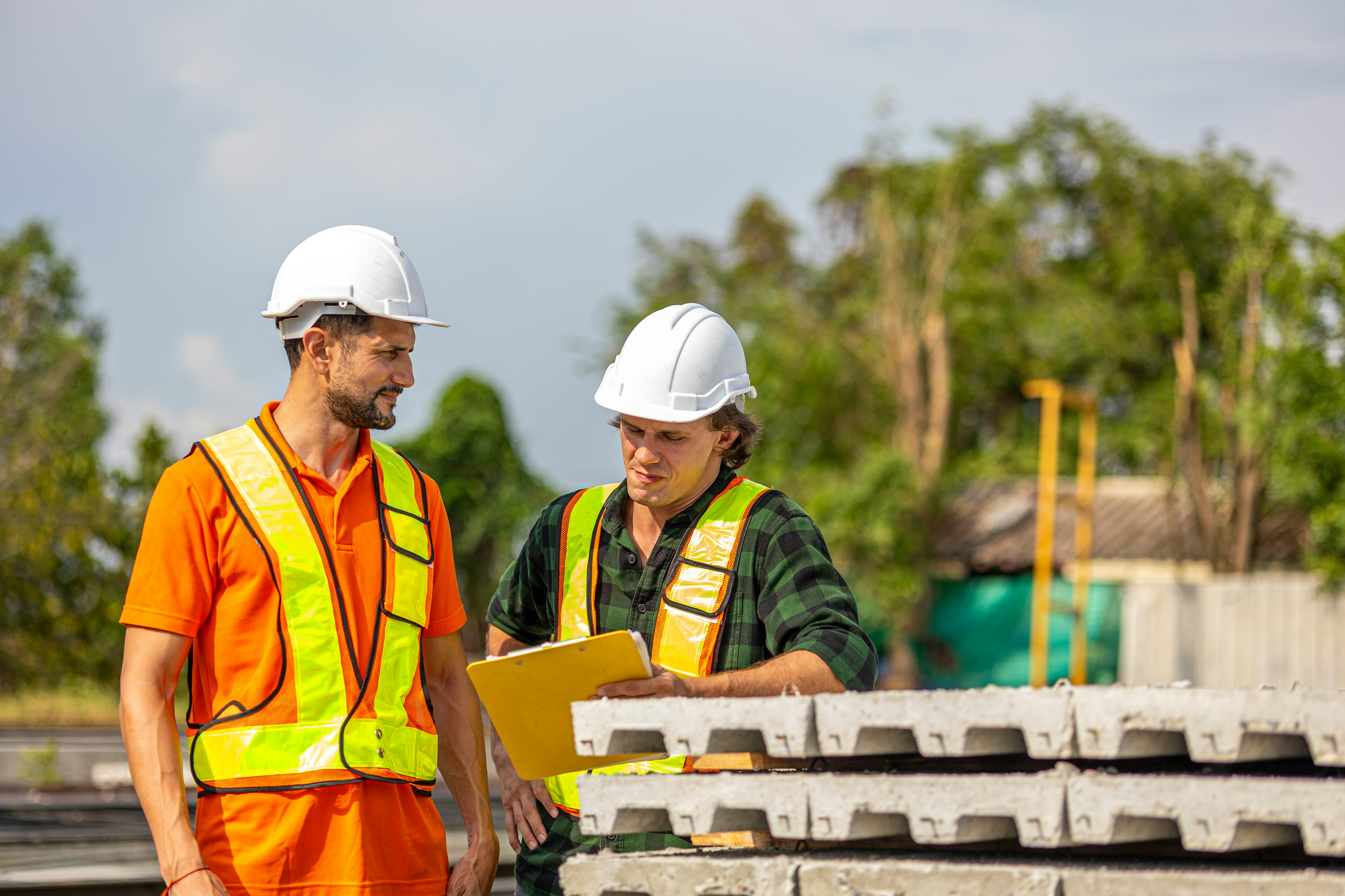 Two site supervisors in hard hats and safety vests reviewing a checklist beside stacked precast concrete panels
