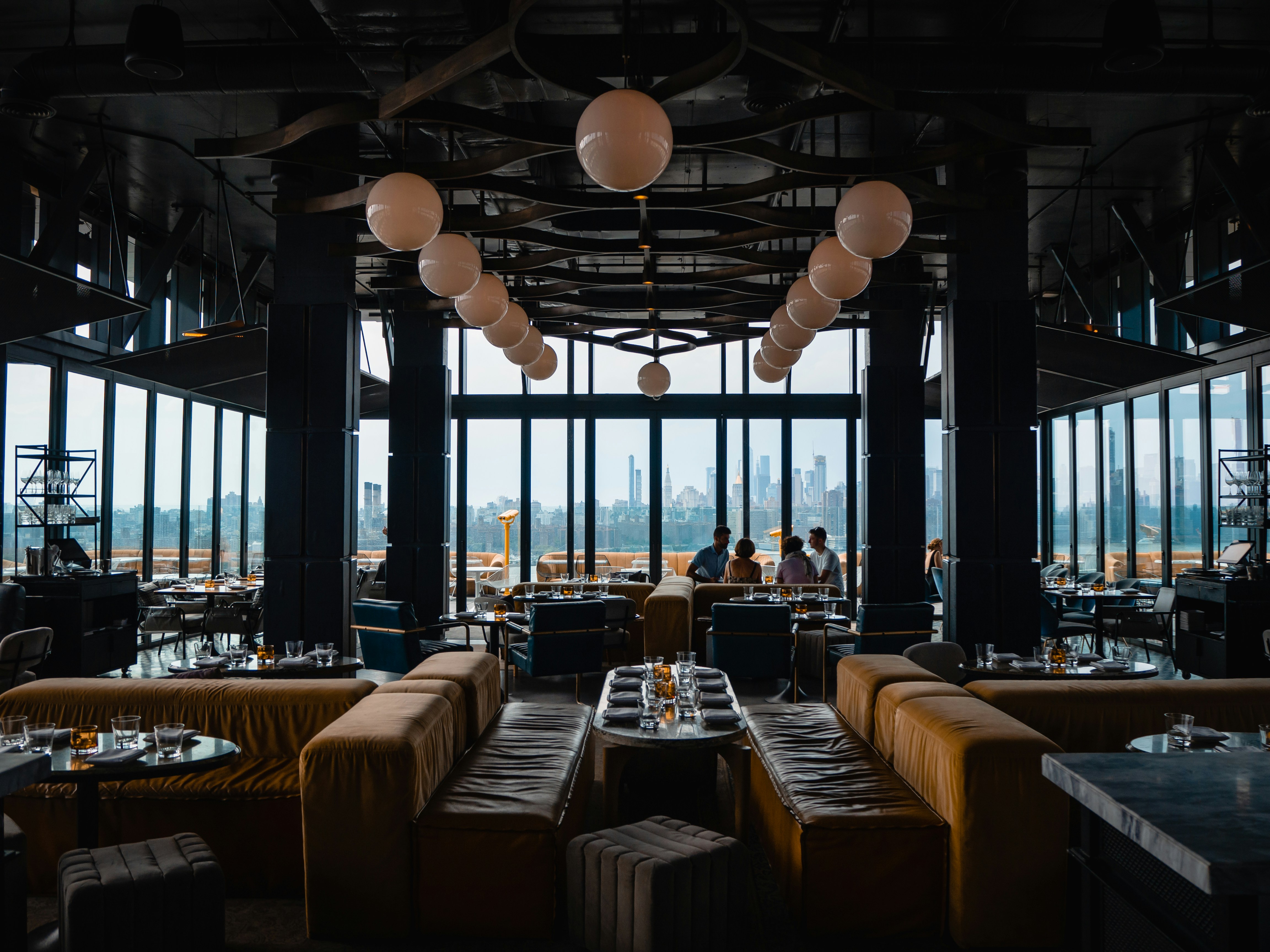 Dark, atmospheric rooftop restaurant interior with velvet banquette seating, globe pendant lighting, and a city skyline view